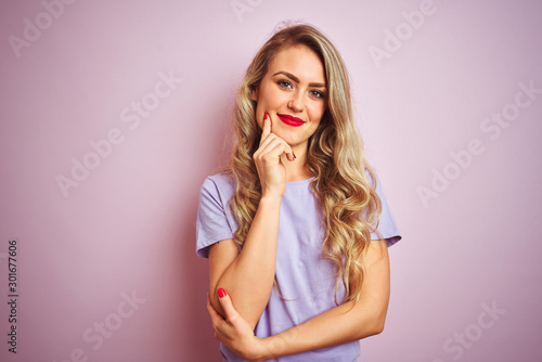 Young beautiful woman wearing purple t-shirt standing over pink isolated background looking confident at the camera smiling with crossed arms and hand raised on chin. Thinking positive.