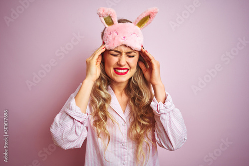 Young beautiful woman wearing pajama and sleep mask over pink isolated background with hand on head for pain in head because stress. Suffering migraine.