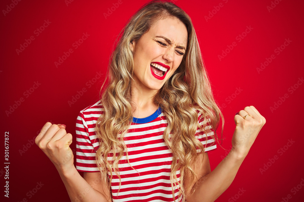 Young beautiful woman wearing stripes t-shirt standing over red isolated background very happy and excited doing winner gesture with arms raised, smiling and screaming for success Young beautiful woman wearing stripes t-shirt standing over red isolated background very happy and excited doing winner gesture with arms raised, smiling and screaming for success