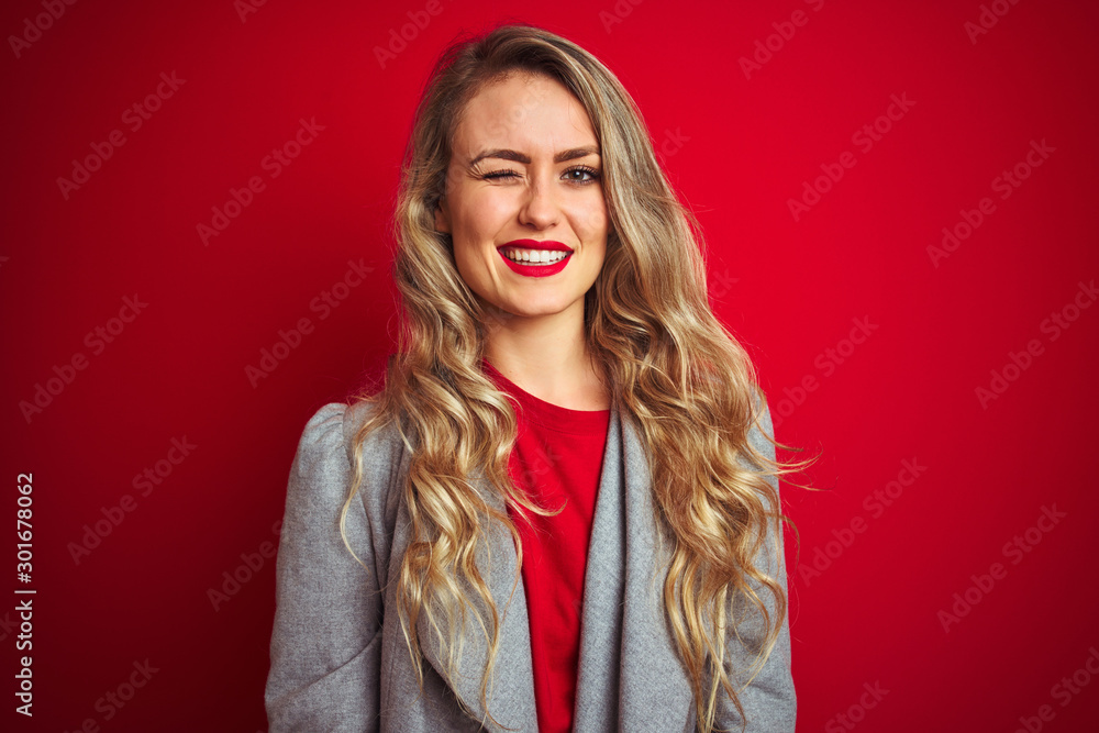 Young beautiful business woman wearing elegant jacket standing over red isolated background winking looking at the camera with sexy expression, cheerful and happy face.
