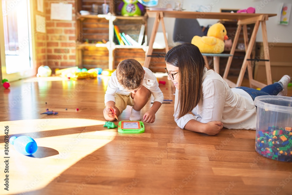 Fototapeta premium Beautiful teacher and toddler drawing using digital board around lots of toys at kindergarten