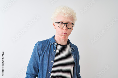 Young albino blond man wearing denim shirt and glasses over isolated white background puffing cheeks with funny face. Mouth inflated with air, crazy expression.