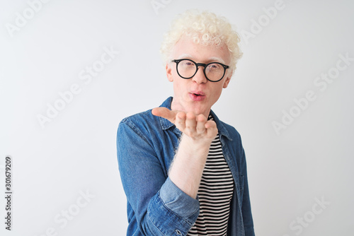Young albino blond man wearing denim shirt and glasses over isolated white background looking at the camera blowing a kiss with hand on air being lovely and sexy. Love expression.