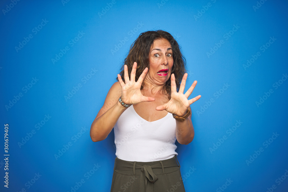 Fototapeta premium Middle age senior woman with curly hair standing over blue isolated background afraid and terrified with fear expression stop gesture with hands, shouting in shock. Panic concept.