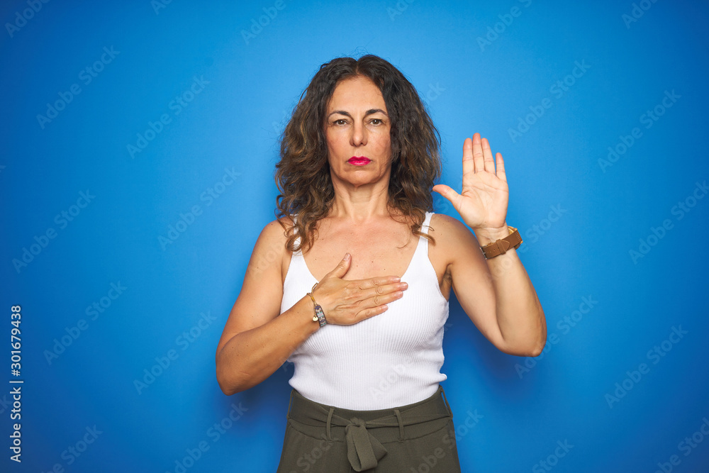 Middle age senior woman with curly hair standing over blue isolated background Swearing with hand on chest and open palm, making a loyalty promise oath Middle age senior woman with curly hair standing over blue isolated background Swearing with hand on chest and open palm, making a loyalty promise oath