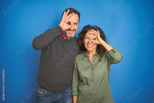 Beautiful middle age couple together standing over isolated blue background doing ok gesture with hand smiling, eye looking through fingers with happy face.