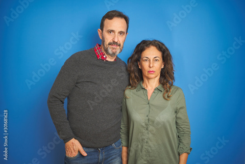 Beautiful middle age couple together standing over isolated blue background with serious expression on face. Simple and natural looking at the camera.