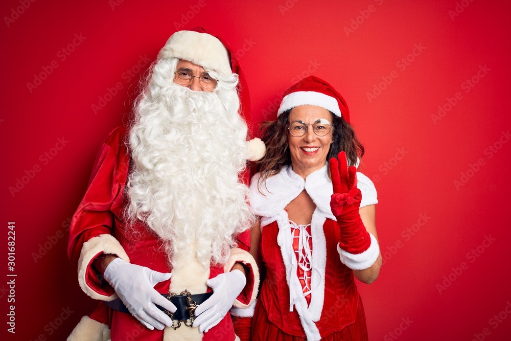 Middle age couple wearing Santa costume and glasses over isolated red background showing and pointing up with fingers number three while smiling confident and happy. Middle age couple wearing Santa costume and glasses over isolated red background showing and pointing up with fingers number three while smiling confident and happy.