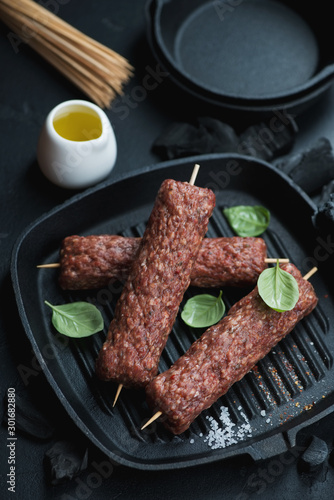 Cast-iron grill pan with fresh uncooked marbled beef kabobs on wooden skewers, studio shot on a black stone surface with charcoals