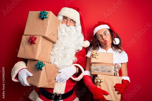 Middle age couple wearing Santa costume holding tower of gifts over isolated red background making fish face with lips, crazy and comical gesture. Funny expression.