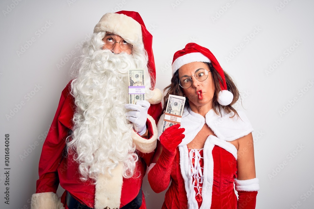Senior couple wearing Santa Claus costume holding dollars over isolated white background making fish face with lips, crazy and comical gesture. Funny expression. Senior couple wearing Santa Claus costume holding dollars over isolated white background making fish face with lips, crazy and comical gesture. Funny expression.