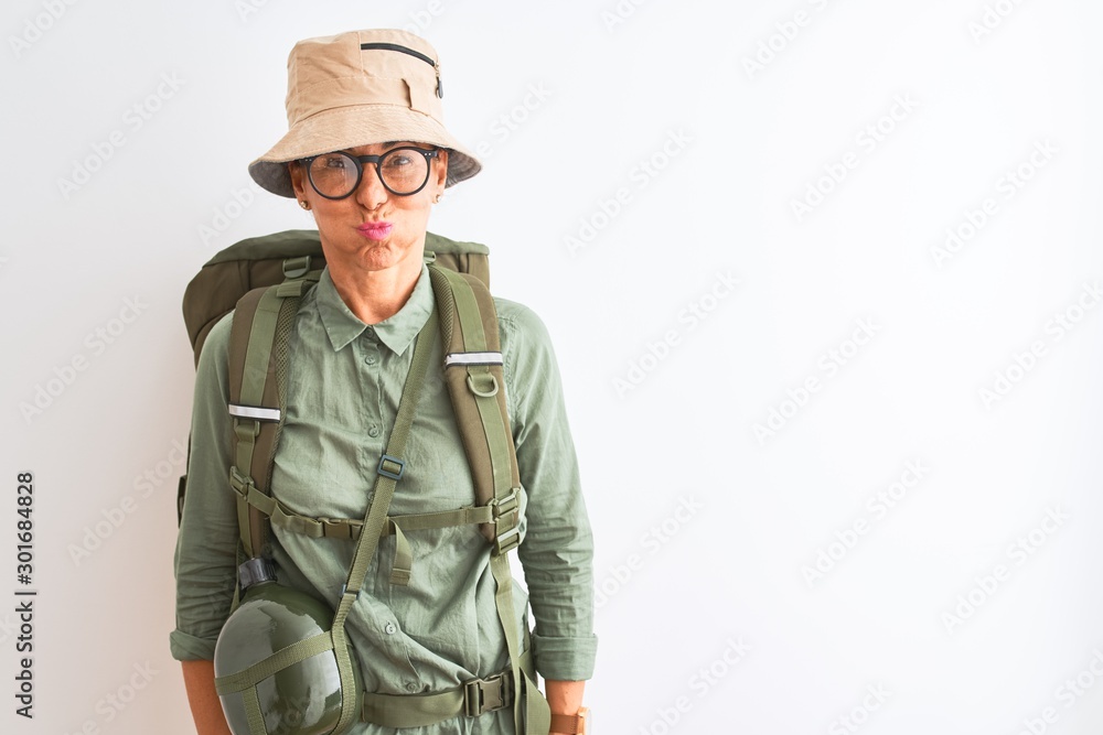 Middle age hiker woman wearing backpack canteen hat glasses over isolated white background puffing cheeks with funny face. Mouth inflated with air, crazy expression.