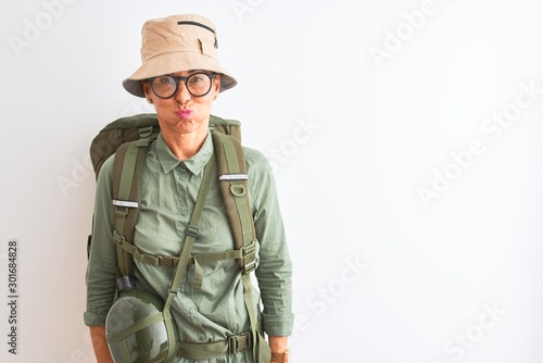 Middle age hiker woman wearing backpack canteen hat glasses over isolated white background puffing cheeks with funny face. Mouth inflated with air, crazy expression.
