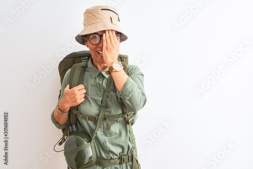 Middle age hiker woman wearing backpack canteen hat glasses over isolated white background covering one eye with hand, confident smile on face and surprise emotion.