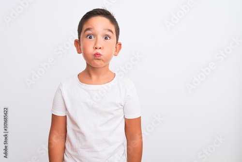 Beautiful kid boy wearing casual t-shirt standing over isolated white background puffing cheeks with funny face. Mouth inflated with air, crazy expression.
