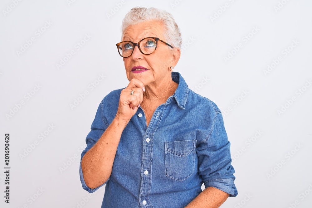 Senior grey-haired woman wearing denim shirt and glasses over isolated white background with hand on chin thinking about question, pensive expression. Smiling and thoughtful face. Doubt concept. Senior grey-haired woman wearing denim shirt and glasses over isolated white background with hand on chin thinking about question, pensive expression. Smiling and thoughtful face. Doubt concept.