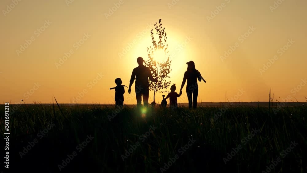 Happy, close-knit family with children, silhouette of farmers outdoors holding hands at sunset. Dad holds a young tree in his hand. The way of life in rural areas. Happy, close-knit family with children, silhouette of farmers outdoors holding hands at sunset. Dad holds a young tree in his hand. The way of life in rural areas.