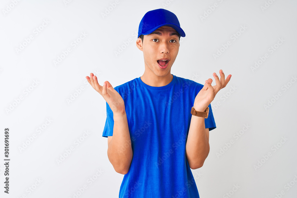 Chinese deliveryman wearing blue t-shirt and cap standing over isolated white background celebrating crazy and amazed for success with arms raised and open eyes screaming excited. Winner concept