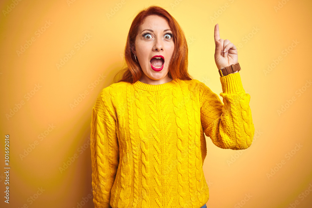 Beautiful redhead woman wearing winter sweater standing over isolated yellow background pointing finger up with successful idea. Exited and happy. Number one. Beautiful redhead woman wearing winter sweater standing over isolated yellow background pointing finger up with successful idea. Exited and happy. Number one.