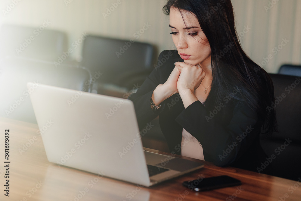 Young attractive emotional girl in business-style clothes sitting at a desk on a laptop and phone in the office or auditorium Young attractive emotional girl in business-style clothes sitting at a desk on a laptop and phone in the office or auditorium