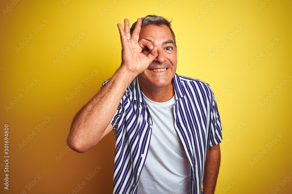 Handsome middle age man wearing striped shirt standing over isolated yellow background doing ok gesture with hand smiling, eye looking through fingers with happy face. Handsome middle age man wearing striped shirt standing over isolated yellow background doing ok gesture with hand smiling, eye looking through fingers with happy face.