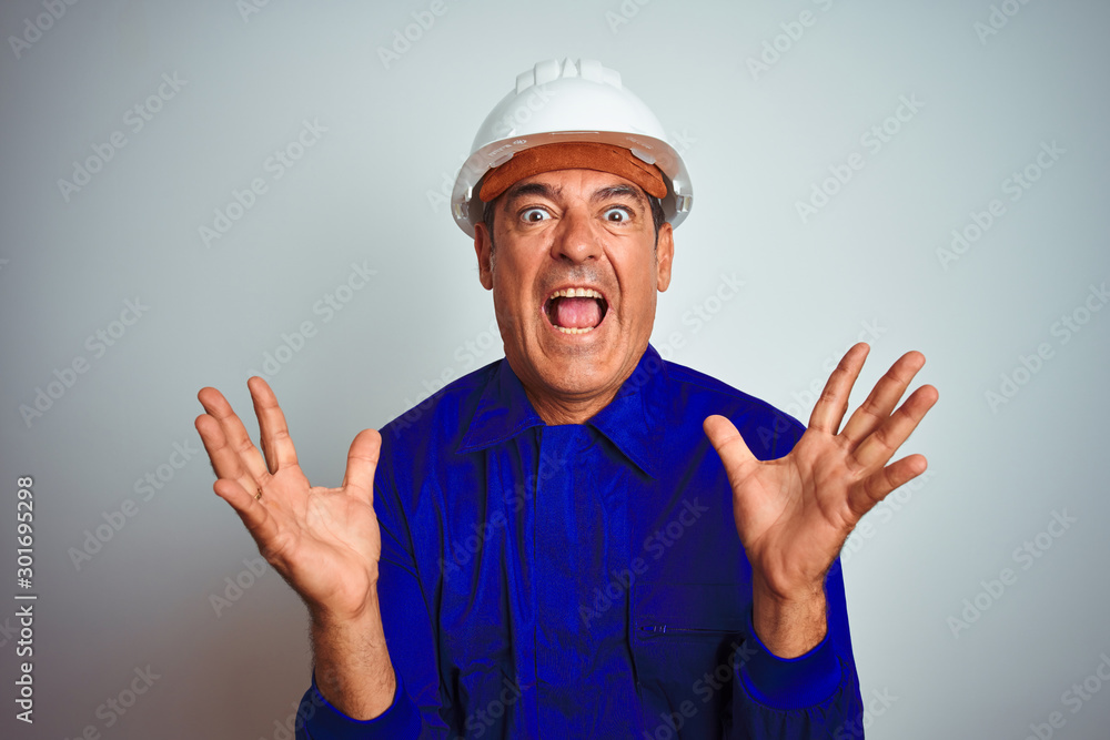 Handsome middle age worker man wearing uniform and helmet over isolated white background crazy and mad shouting and yelling with aggressive expression and arms raised. Frustration concept.
