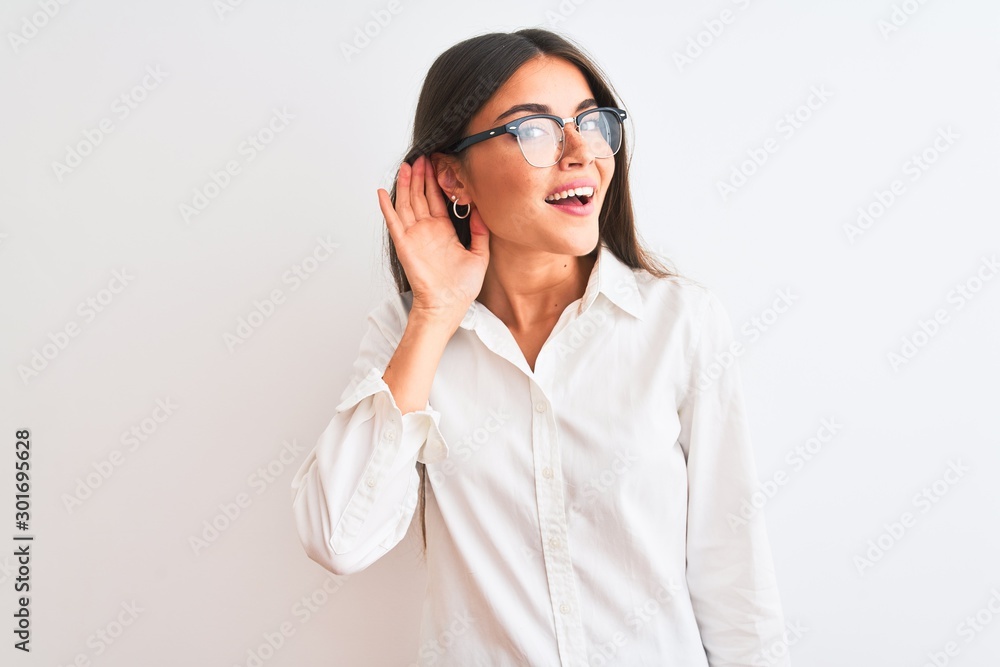 Young beautiful businesswoman wearing glasses standing over isolated white background smiling with hand over ear listening an hearing to rumor or gossip. Deafness concept.
