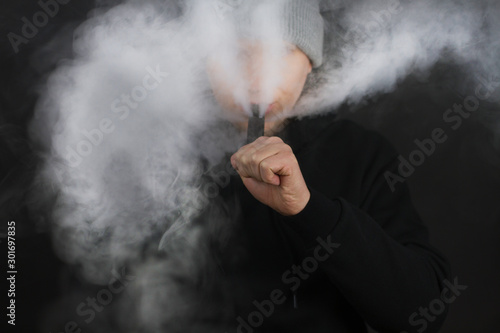 The man smoke an electronic cigarette on the dark background, Men in hoody vaping and releases a cloud of vapor. Guy with vaping on black background © paulcannoby