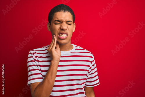 Young handsome arab man wearing striped t-shirt over isolated red background touching mouth with hand with painful expression because of toothache or dental illness on teeth. Dentist concept.