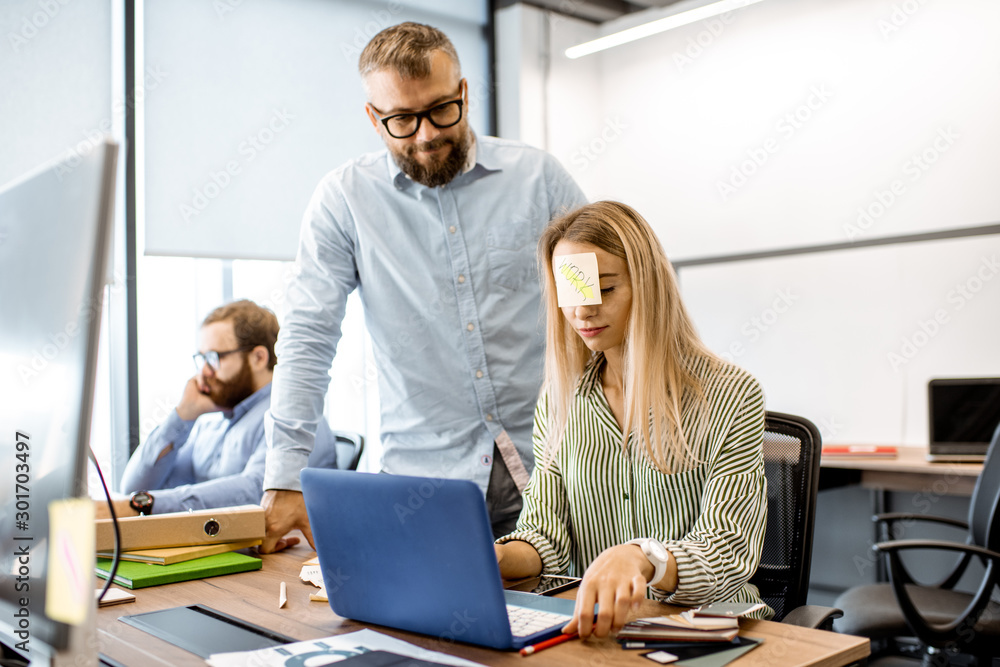 Young woman with work reminder sticker on her head feeling tired during the hard work with coworkers in the office Young woman with work reminder sticker on her head feeling tired during the hard work with coworkers in the office