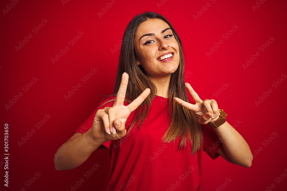 Young beautiful woman wearing t-shirt standing over isolated red background smiling looking to the camera showing fingers doing victory sign. Number two. Young beautiful woman wearing t-shirt standing over isolated red background smiling looking to the camera showing fingers doing victory sign. Number two.