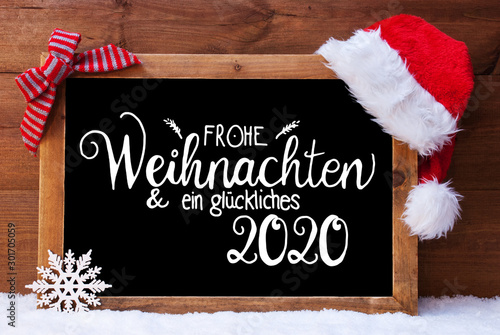 Chalkboard With German Calligraphy Frohe Weihnachten Und Ein Glueckliches 2020 Means Merry Christmas And A Happy 2020. Christmas Decoration Like Santa Hat And Bow. Wooden Background With Snow