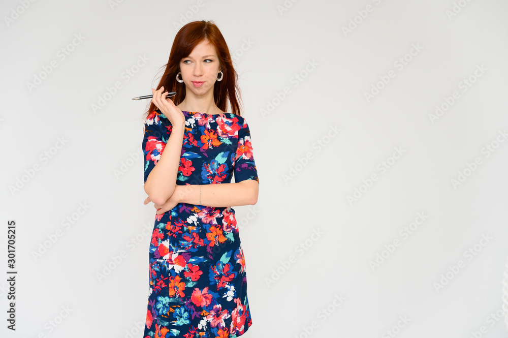 Portrait of a pretty woman, red-haired student girl with long straight hair on a white background in a multi-colored dress with a pen in her hands. Talking to the camera, showing emotions, smiling. Portrait of a pretty woman, red-haired student girl with long straight hair on a white background in a multi-colored dress with a pen in her hands. Talking to the camera, showing emotions, smiling.