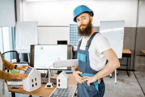 Portrait of a bearded worker in workwear and protective helmet standing in the alternative energy design office