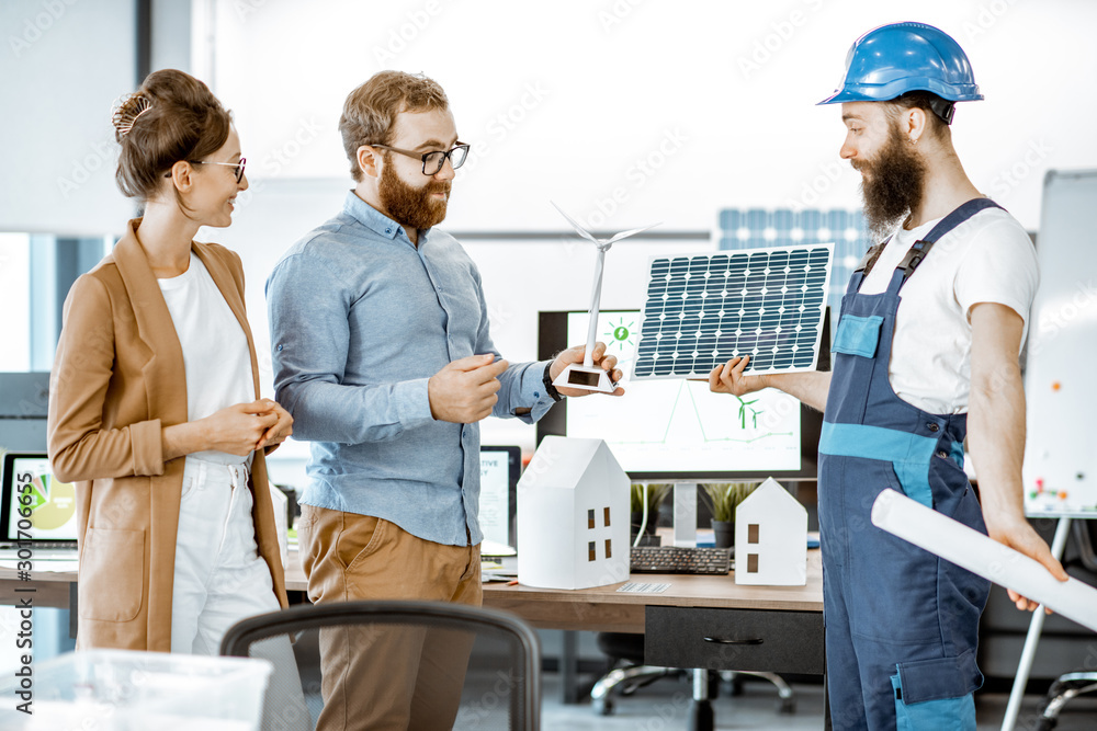 Young engineers and workman working on a project of alternative energy, holding house and wind turbine model in the office Young engineers and workman working on a project of alternative energy, holding house and wind turbine model in the office