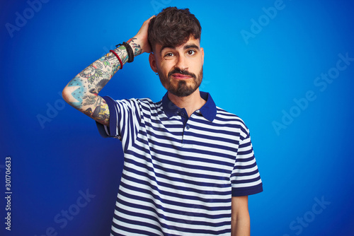 Young man with tattoo wearing striped polo standing over isolated blue background confuse and wonder about question. Uncertain with doubt, thinking with hand on head. Pensive concept.