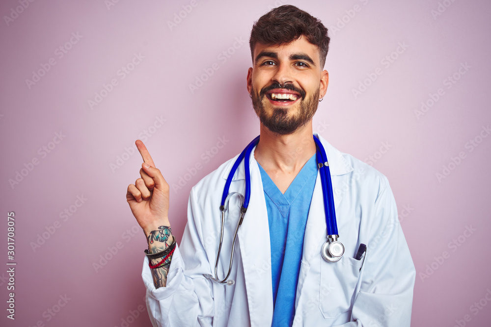 Young doctor man with tattoo wearing stethocope standing over isolated pink background with a big smile on face, pointing with hand and finger to the side looking at the camera. Young doctor man with tattoo wearing stethocope standing over isolated pink background with a big smile on face, pointing with hand and finger to the side looking at the camera.