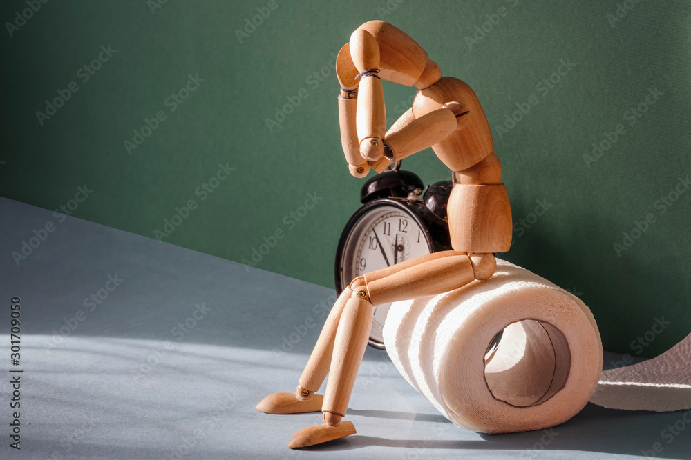 Wooden figure sit on a roll of toilet paper near alarm clock. Concept of the problem with digestion.