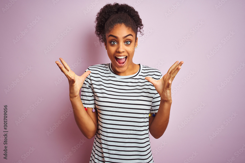African american woman wearing navy striped t-shirt standing over isolated pink background celebrating crazy and amazed for success with arms raised and open eyes screaming excited. Winner concept African american woman wearing navy striped t-shirt standing over isolated pink background celebrating crazy and amazed for success with arms raised and open eyes screaming excited. Winner concept
