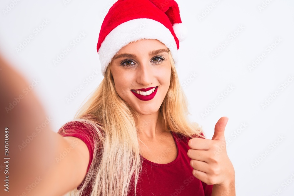 Woman wearing Christmas Santa hat make selfie by camera over isolated white background happy with big smile doing ok sign, thumb up with fingers, excellent sign Woman wearing Christmas Santa hat make selfie by camera over isolated white background happy with big smile doing ok sign, thumb up with fingers, excellent sign