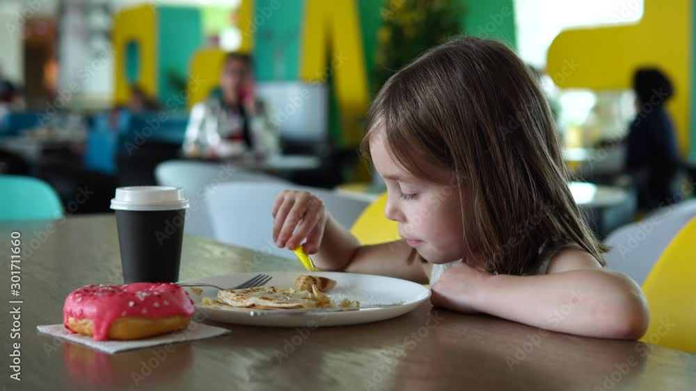One child sits at a table in a children's cafe. A girl of Russian appearance, 5-6 years old. She is eating a donut and pancake. One child sits at a table in a children's cafe. A girl of Russian appearance, 5-6 years old. She is eating a donut and pancake.