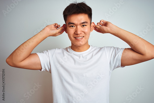 Young asian chinese man wearing t-shirt standing over isolated white background Smiling pulling ears with fingers, funny gesture. Audition problem