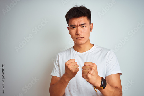 Young asian chinese man wearing t-shirt standing over isolated white background Ready to fight with fist defense gesture, angry and upset face, afraid of problem