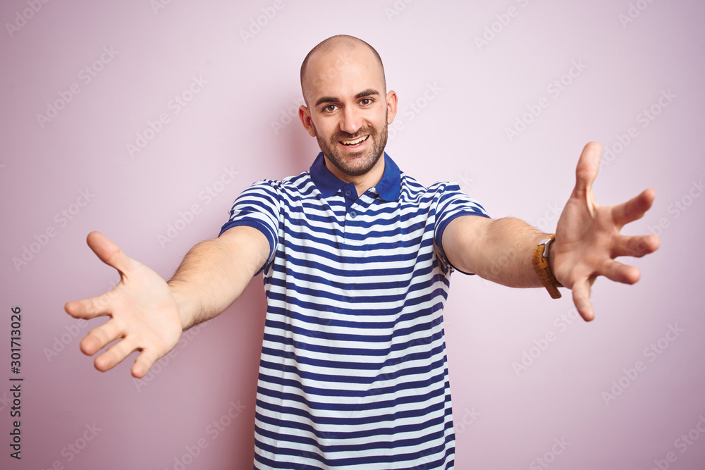Young bald man with beard wearing casual striped blue t-shirt over pink isolated background looking at the camera smiling with open arms for hug. Cheerful expression embracing happiness. Young bald man with beard wearing casual striped blue t-shirt over pink isolated background looking at the camera smiling with open arms for hug. Cheerful expression embracing happiness.