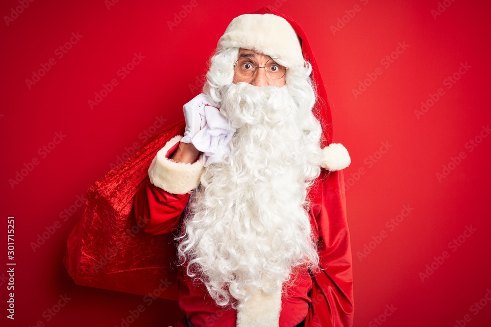 Middle age man wearing Santa costume holding sack with gifts over isolated red background scared in shock with a surprise face, afraid and excited with fear expression