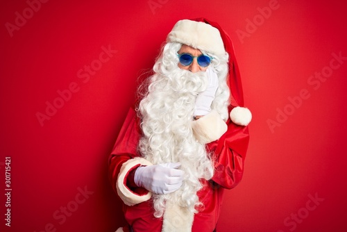Middle age man wearing Santa Claus costume and sunglasses over isolated red background thinking looking tired and bored with depression problems with crossed arms.