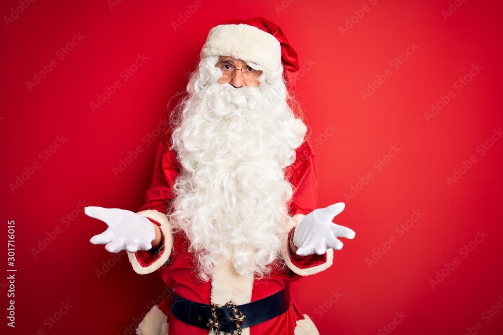 Middle age handsome man wearing Santa costume standing over isolated red background clueless and confused with open arms, no idea concept.
