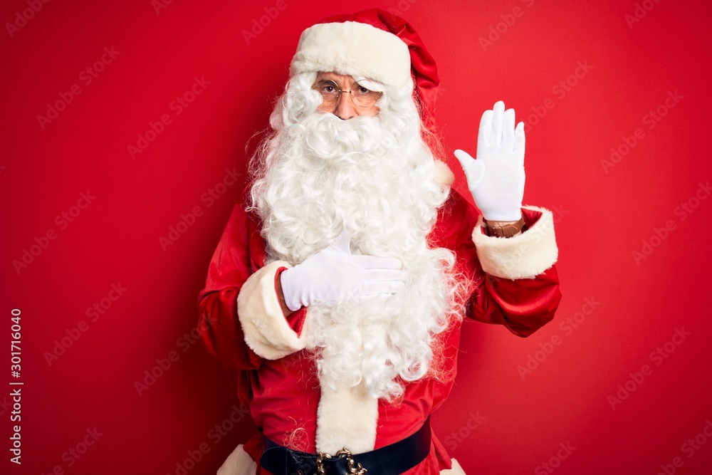 Middle age handsome man wearing Santa costume standing over isolated red background Swearing with hand on chest and open palm, making a loyalty promise oath