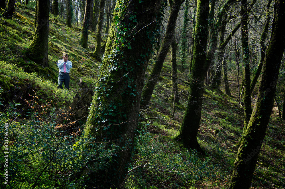 Lost businessman in shirt and tie standing outdoors on a wooded hillside surrounded by a forest of green trees Lost businessman in shirt and tie standing outdoors on a wooded hillside surrounded by a forest of green trees