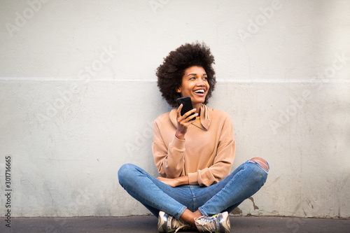 happy young african american woman with afro hair sitting on floor with cellphone